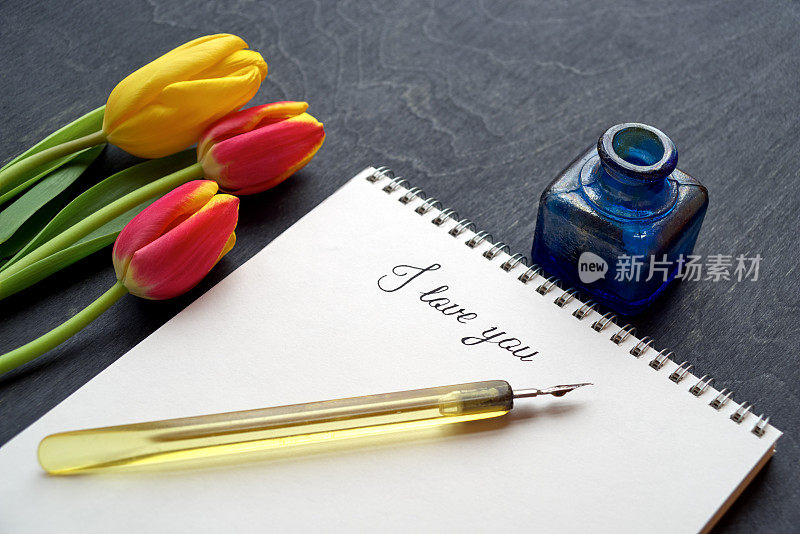 旧钢笔和词“我爱你”在纸笔记本上的黑色背景与鲜花和墨水瓶