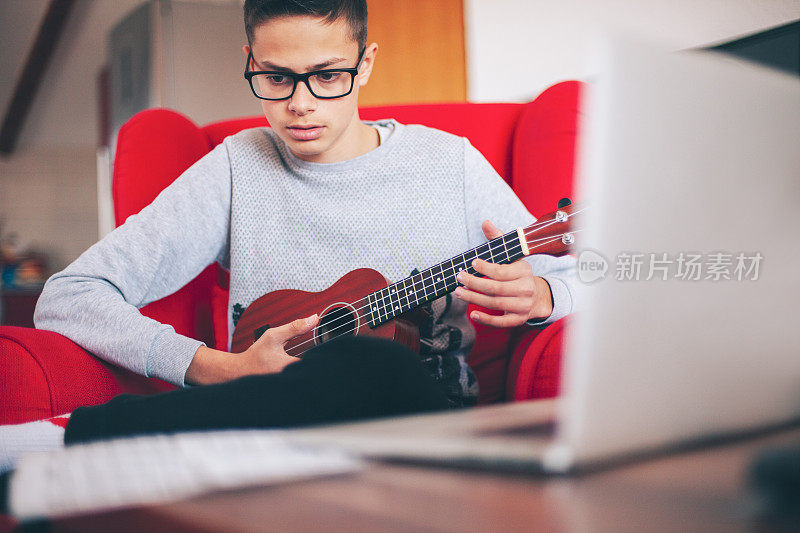 由于冠状病毒隔离，坐在红色扶手椅上的孩子在隔离期间学习弹奏尤克里里琴