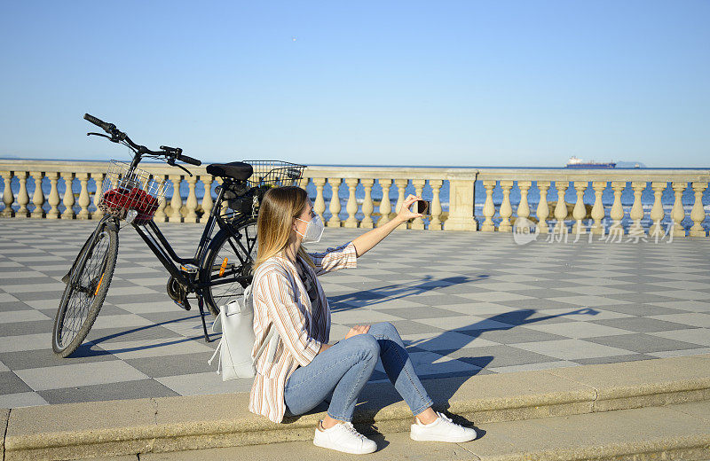 年轻女子戴着防护面具骑着电动自行车在海边使用手机