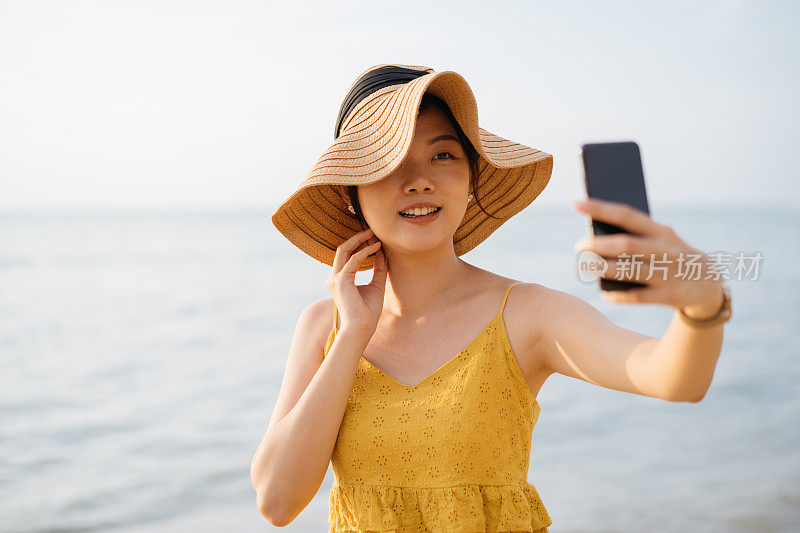 年轻的亚洲女游客戴着太阳帽用智能手机在海滩自拍