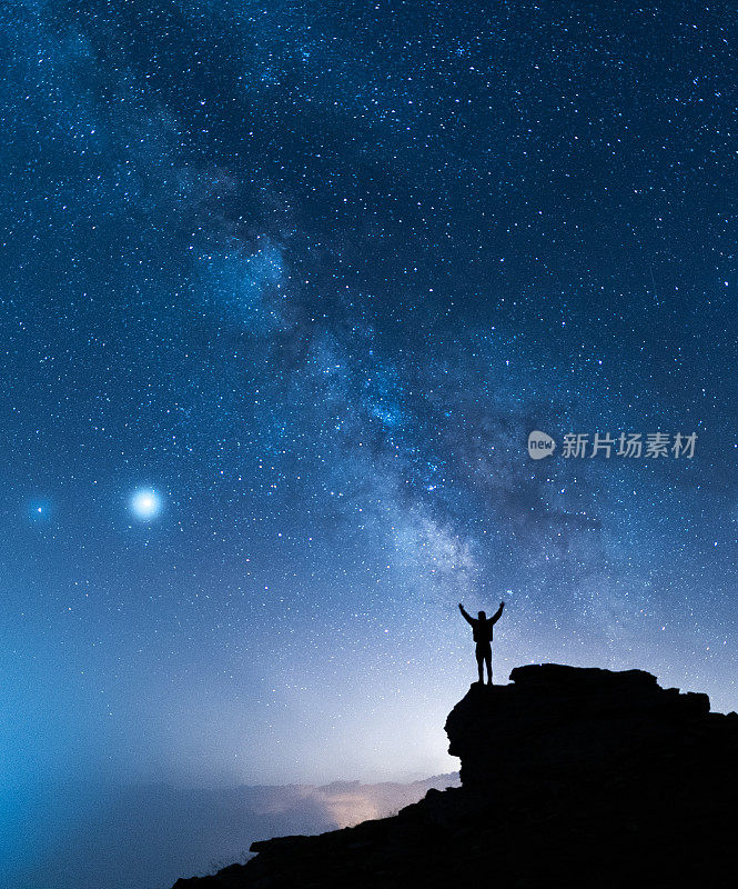 人与宇宙。一个人站在银河系旁边的山顶上，双手举向空中。