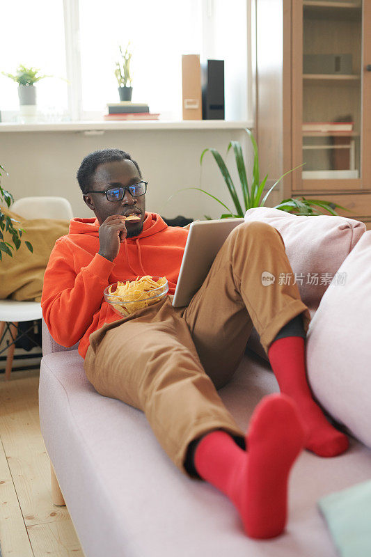 英俊的非裔美国人穿着休闲衣服躺在沙发上吃薯片，在数字平板电脑上看电视剧的垂直高角度拍摄