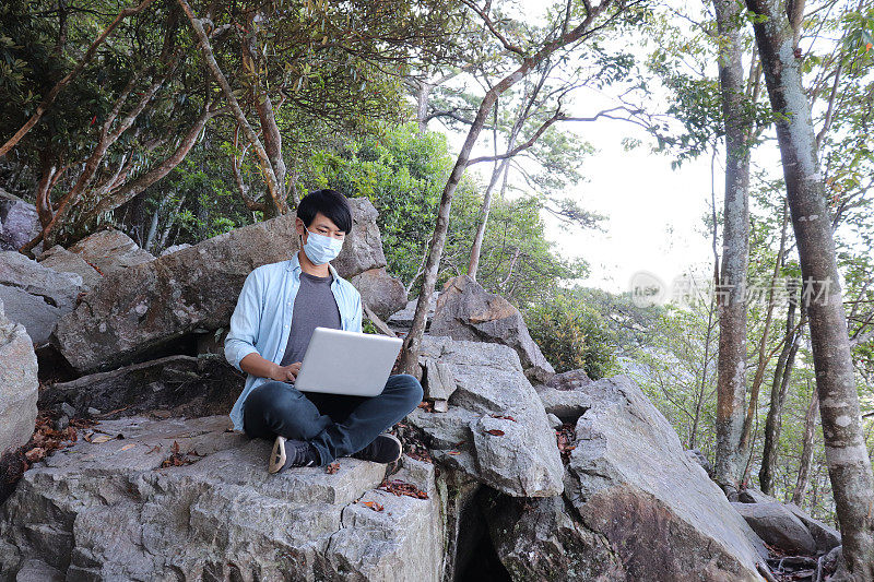 亚洲人戴着面具，用笔记本在山上工作。