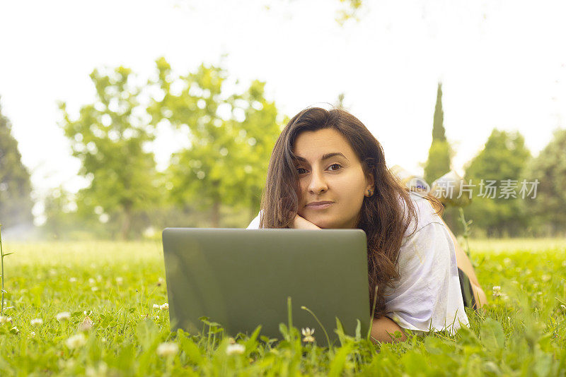 年轻女子在户外用笔记本电脑工作，手放在下巴上看着相机，微笑着。
