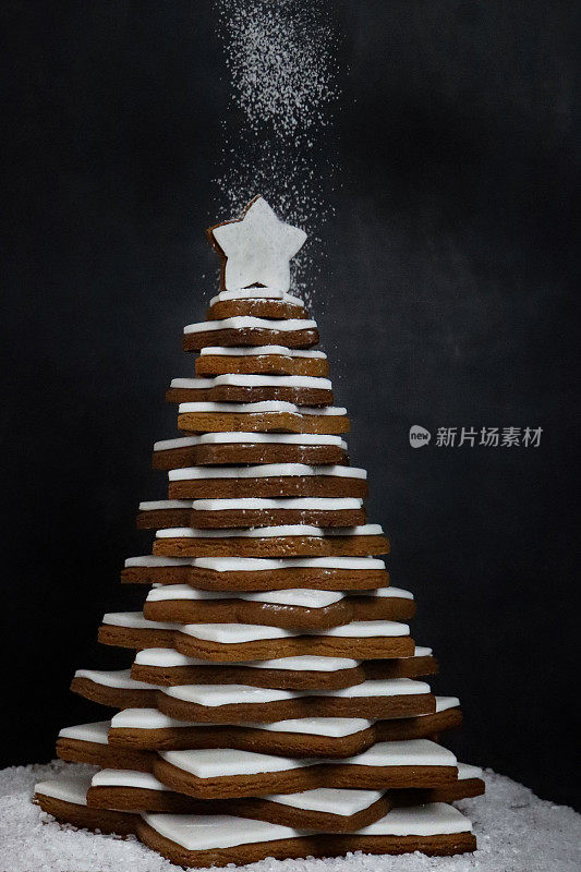 糖霜被筛选在圣诞树上的图像，圣诞树是由堆叠的姜饼星饼干形成的，上面覆盖着白色的皇家糖霜，人造雪，黑色的背景，集中在前景上