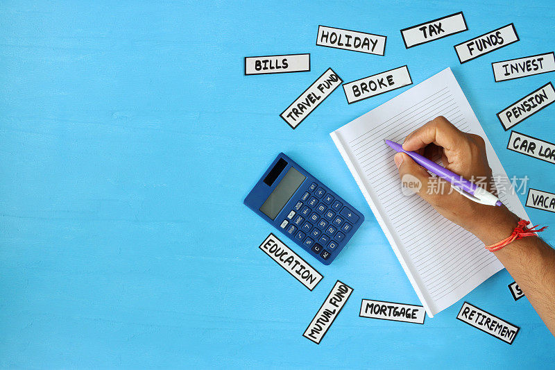 图片中的人正在使用横格纸，计算器和笔，周围是标有财务单词的标签，蓝色背景，高架视野，家庭财务和储蓄概念，复印空间
