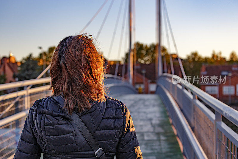 后视图的红发女人走在桥在日落。背景是住宅区。Alcorcon,马德里,西班牙。