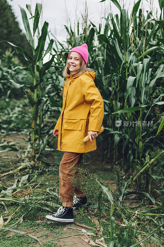 一个穿着黄色雨衣，戴着一顶粉红色帽子的时髦少女在一片玉米地上跳舞和大笑