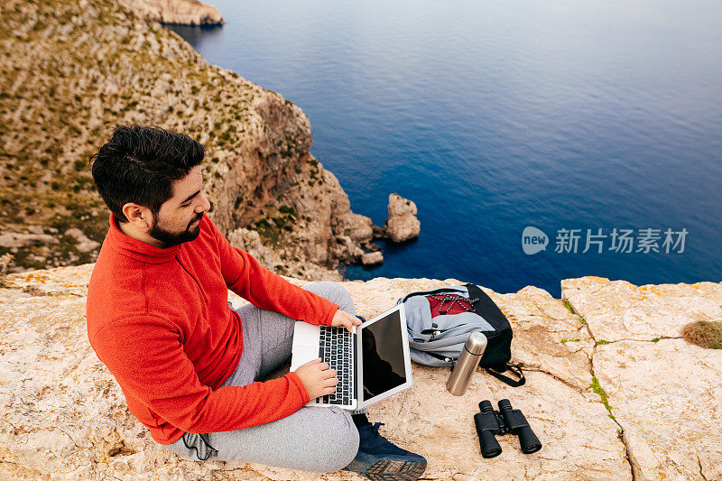 身穿红色运动衫的年轻徒步旅行者，拿着打开的笔记本电脑坐在一堵岩石墙上，背景是马略卡岛北部海岸的波伦萨湾