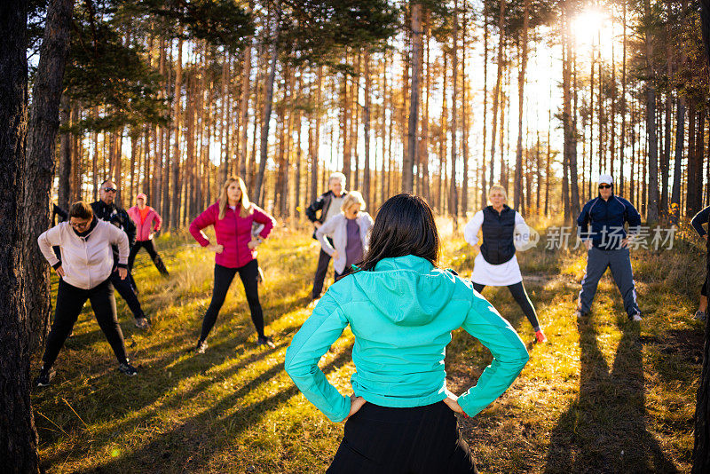 在森林里，一群白种人在教练的带领下进行集体健身课程，伸展身体