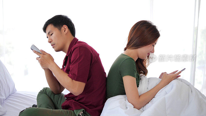 一对亚洲情侣坐在床上，用手机发短信，背对着对方，用智能手机。冷漠与不重视彼此