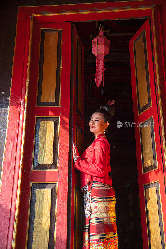 近距离观察身穿泰国兰纳传统服饰的年轻亚洲女子，用红色站在门口，古屋的古风也面带微笑地望向左边。