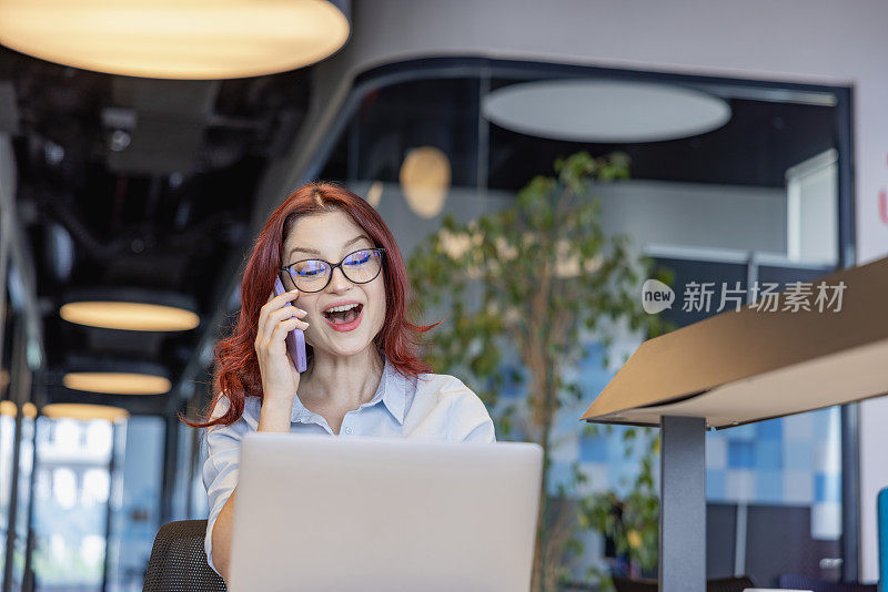 一个年轻迷人的女商人的肖像，在一个开放的现代办公室里，一边使用笔记本电脑和打电话，一边享受工作。