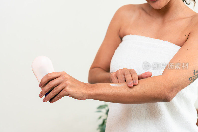 未被认出的女士沐浴后用白色毛巾在手臂上涂抹新的活肤美容产品，改善保湿和柔软的皮肤。拉丁女性每天早晨在卧室里做例行美容程序，在手臂上涂乳液。