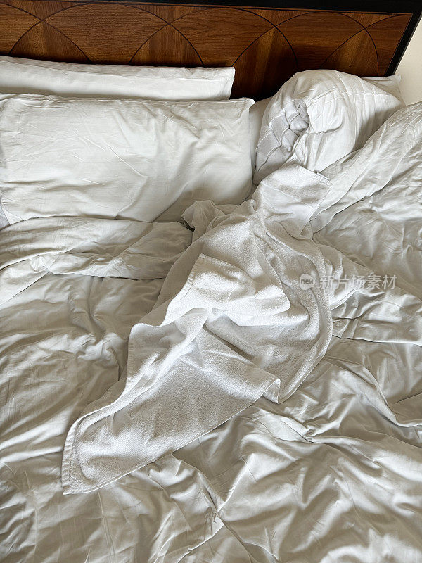 酒店卧室里凌乱的双人床、木制床头板、褶皱的床单、褶皱的枕头和羽绒被的全画幅图片