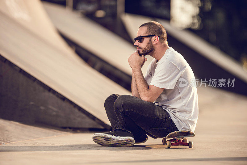 在滑板公园里，一个沉思的年轻人坐在滑板上。