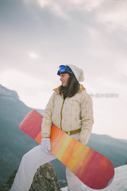 女人拿着滑雪板，背景是高山