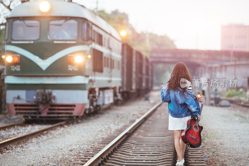 美丽的女孩拿着吉他走在火车站的铁轨上