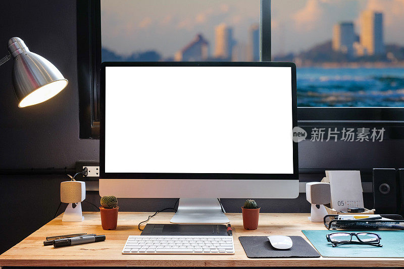 平面设计师的工作空间配备了一个手写板，一台电脑和白色的文本屏幕与美丽的日落从窗口的海滩