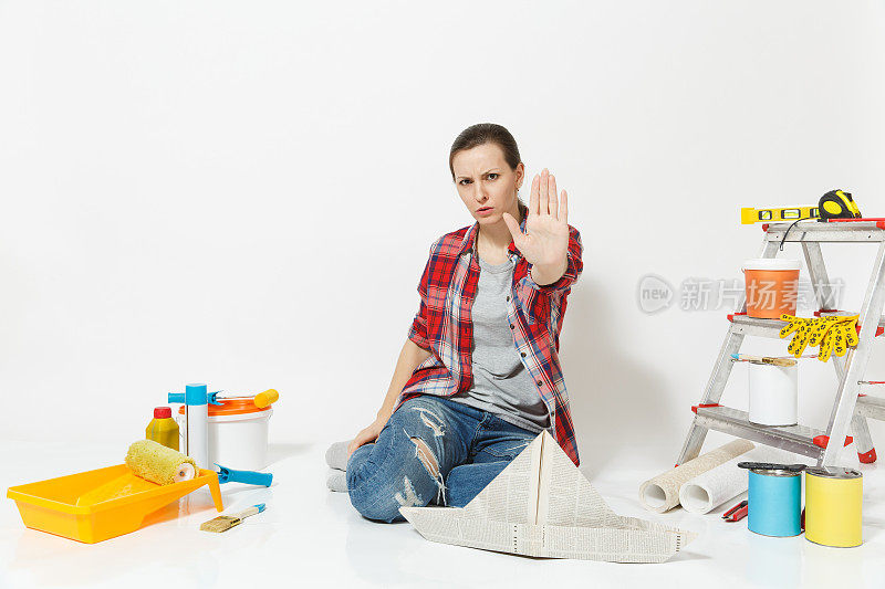 严厉的女人坐在地板上戴着报纸帽，仪器为装修公寓房间，显示停止手势与手掌孤立在白色的背景。壁纸、配件。修理家里的概念。
