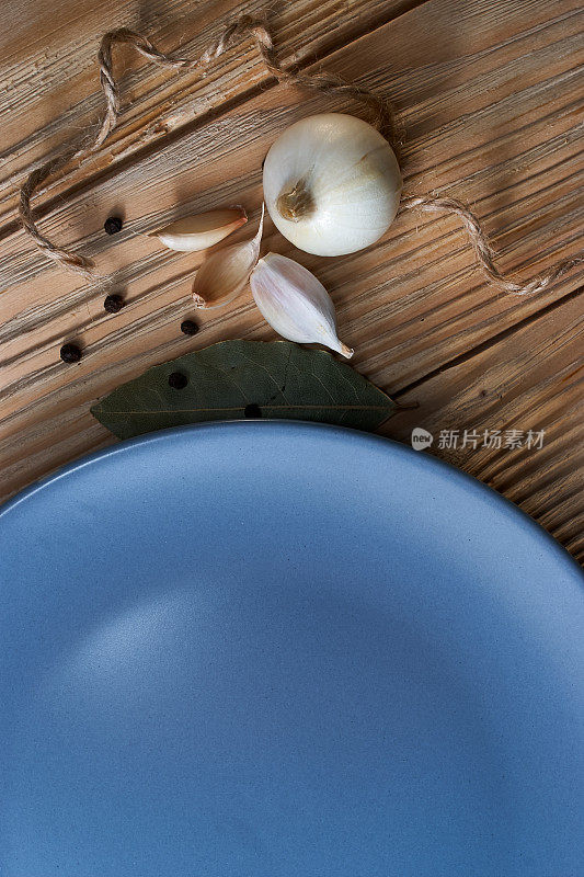 木桌面上放着月桂叶、洋葱、大蒜和空的蓝色盘子。干香料混合