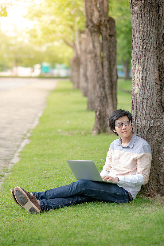 年轻的亚洲男子大学学生戴眼镜坐在绿色草地上使用笔记本电脑在大学公园。高中的校园生活。教育机会或奖学金概念
