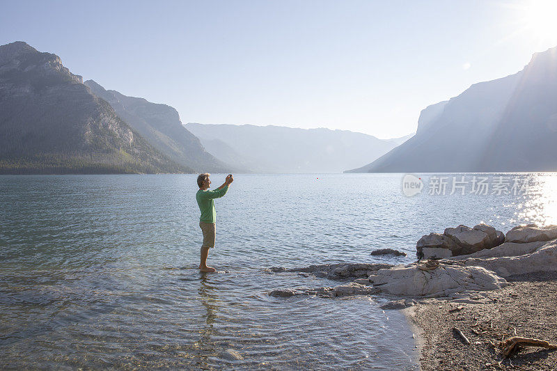 一名男性徒步旅行者在日出时的湖底用智能手机拍照