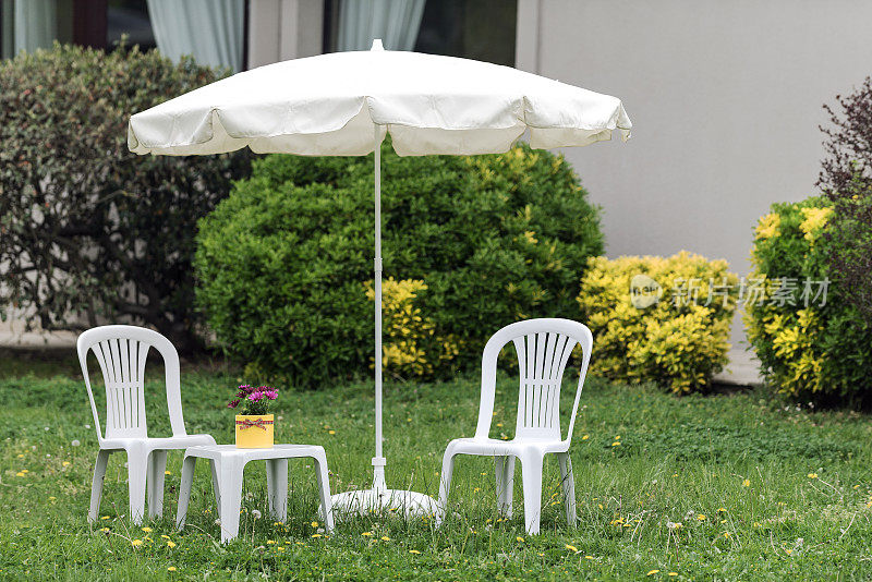白色太阳伞，塑料椅子和咖啡桌在花园里的草地上
