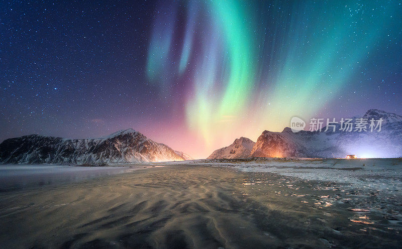 冬天的北极光出现在雪山和沙滩上。挪威罗浮敦群岛的北极光。极光点缀着星空。夜景有极光，冰冻的海岸，城市灯光