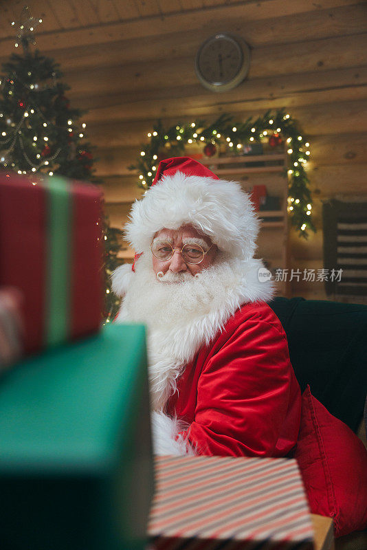 圣诞老人从一堆礼品盒后面看着我们，坐在他的书房里，放着圣诞装饰品和圣诞树，