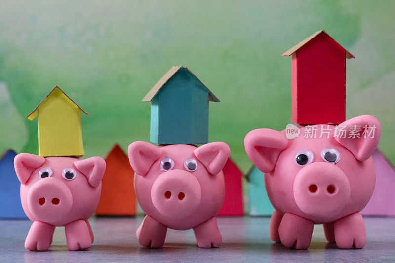 三只模型猪的特写图像与模型纸板房子平衡在小猪存钱罐的背部，排彩色的海滩小屋，重点在前景，斑白的绿色背景，家庭财务，房地产和假日储蓄概念，复制空间