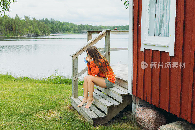 在瑞典，一名女性凝视着夏天的早晨，在湖边的红色小屋旁喝着咖啡