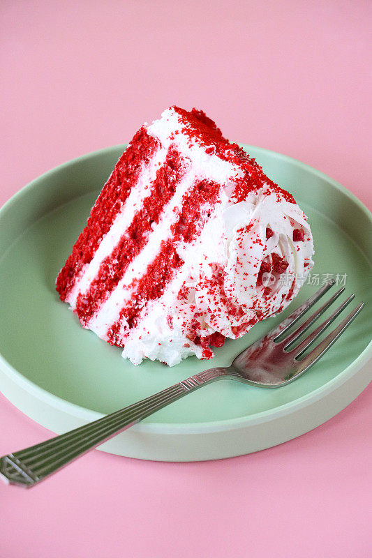 红色天鹅绒蛋糕片的图像，三层蛋糕被奶油在绿色盘子上用叉子分开，管道糖衣玫瑰，脱水覆盆子粉，粉色背景，重点在前景