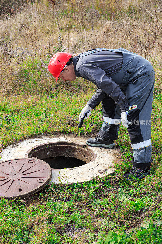一名穿着工作服的男性管道工用手电筒照着水井。检查和修理管道系统