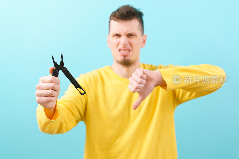 一个男人拿着一个多功能工具和钳子在一个设备上，并在蓝色背景上展示他的拇指。口袋里。黑色的。刀片。设备。金属。多用途。实用程序。生存。开放。折叠。对象。处理