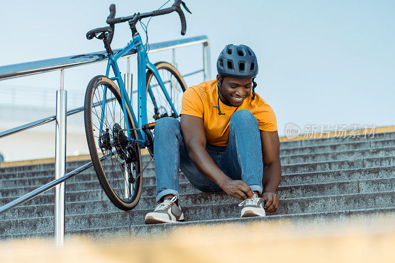 微笑的尼日利亚男子戴着安全帽，坐在楼梯上系鞋带，靠近自行车复印处