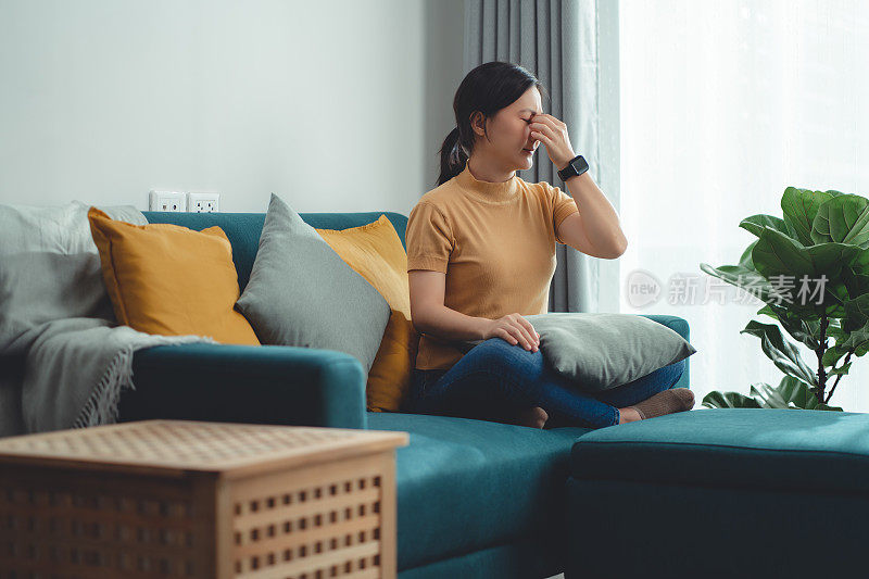 一名亚洲妇女坐在家里客厅的沙发上遭受眼睛疼痛。