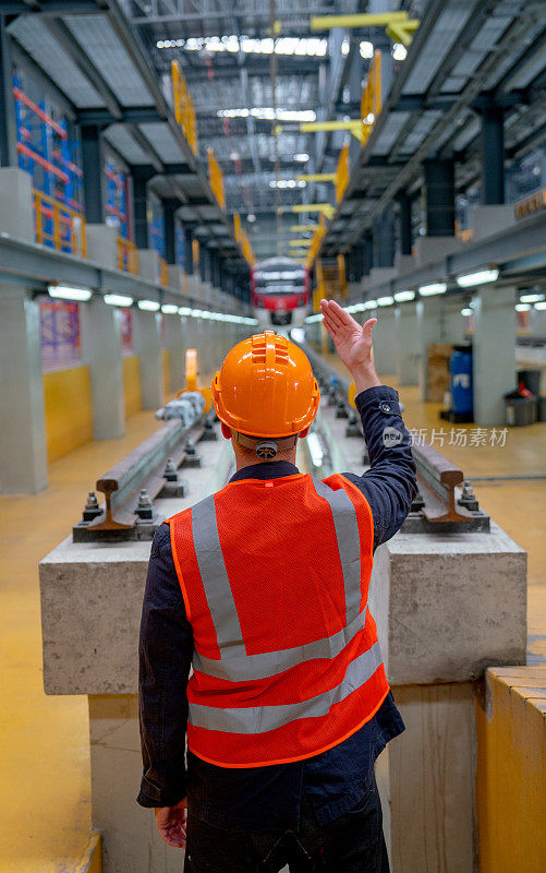 穿着安全制服的技术工人的背部垂直图像，向电气或地铁列车显示身体信号，并站在工厂工作场所的铁轨前。