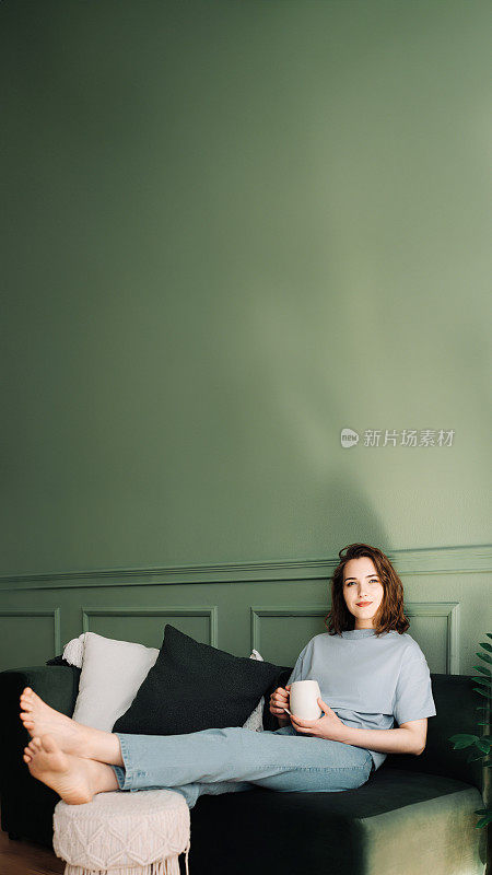 一位若有所思的年轻中年妇女在沙发上享受着休息和一杯茶，沉思着，看着开阔的空间