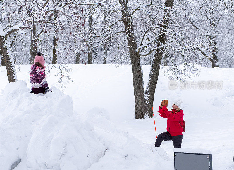 一位年轻的白人妇女用智能手机给一个孩子拍照;孩子坐在一大堆雪里微笑着。