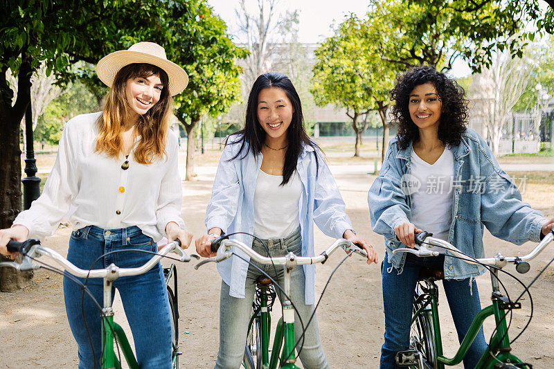 三位千禧一代的年轻女性朋友站在户外，骑着自行车