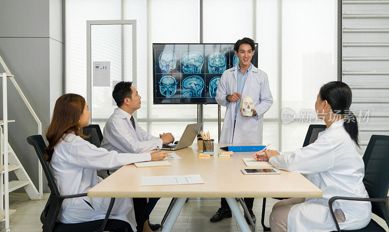 年轻的医学专业人员站着展示，拿着一个人类头骨模型，参考屏幕上显示的大脑扫描图像。听众聚精会神地听讲演。