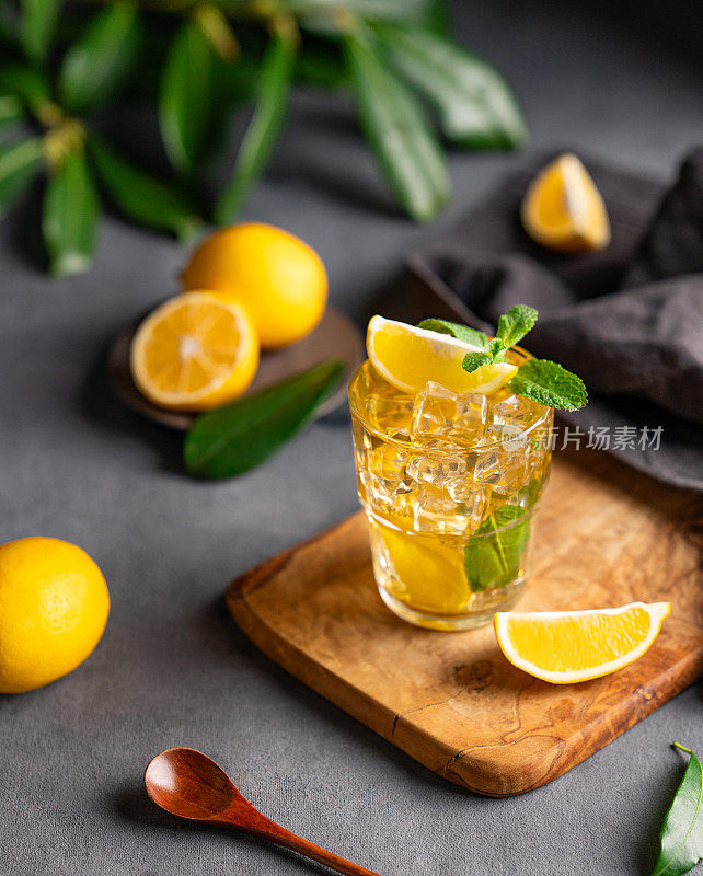 一杯柠檬和薄荷冰茶，放在木板上，深色背景上点缀着柑橘类水果和树枝。