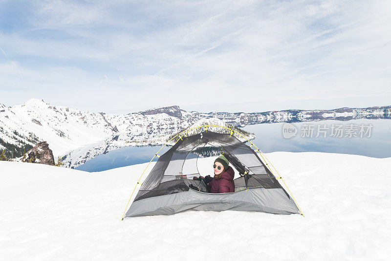 一位女性徒步旅行者在她舒适的帐篷里放松，帐篷搭在雪地上俯瞰火山口湖