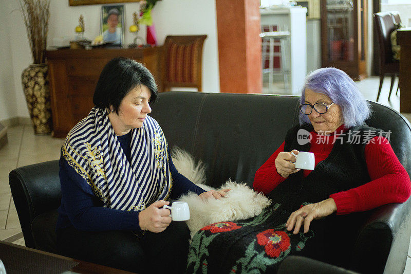 漂亮的老妈妈和她的成年女儿拿着杯子，坐在家里的沙发上聊天