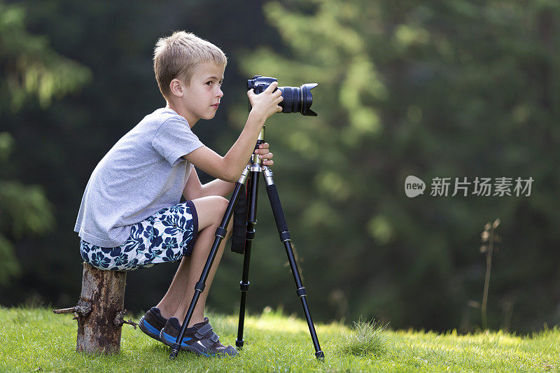 金发小男孩坐在树桩上的草地上用三脚架相机拍照。