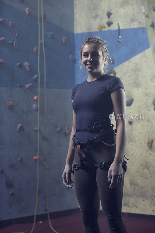 年轻的女学生带着登山装备微笑地站在攀岩墙前