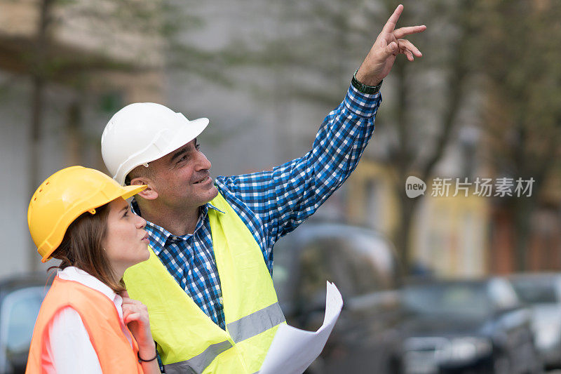 两名建筑工人，一男一女，穿着橙色和黄色的安全夹克和头盔指着他们上方的什么东西