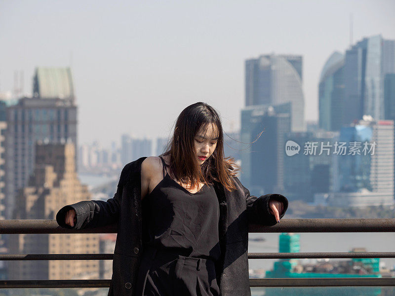 身着黑色外套的黑发美女站在大厦屋顶上，以上海外滩标志性建筑为背景，低头迎风。情感、人、美、生活理念。