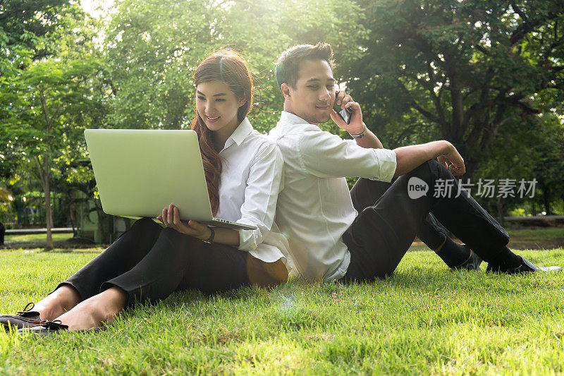 快乐的亚洲夫妇大学工作与科技笔记本电脑和通过智能手机在公园与镜头光晕，商业和生活方式的概念